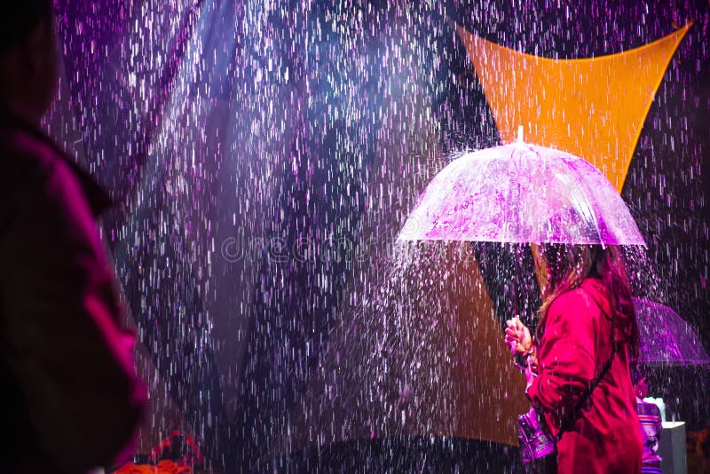 Back view female person with umbrella under artificial rain coming from the ceiling and illuminated by colored lights. Unusual. Event entertainment solution stock photography