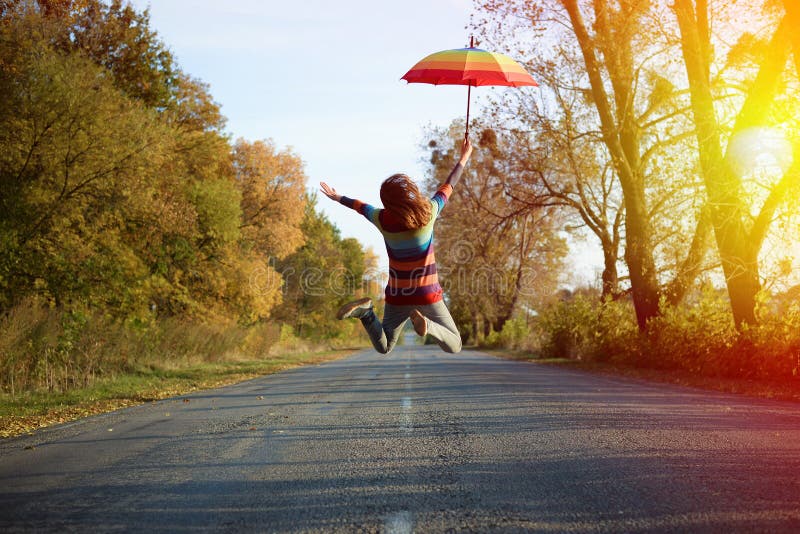Picture of jumping lady holding umbrella with arms. Conceptual picture of jumping lady holding umbrella with arms sideways on empty country road in autumn stock photo