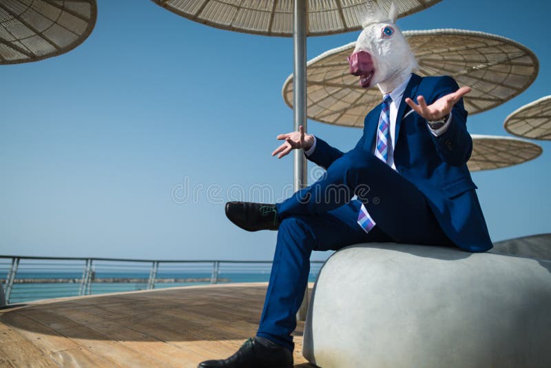 Young businessman in suit sits under umbrellas on the city waterfront. And spreads his hands. Unusual man in funny mask gesticulate on city promenade. Elegant stock images