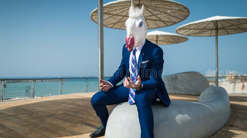 Young elegant man in suit sits under umbrellas on the city waterfront. And active gesticulate. Unusual traveler in comical mask spreads his hands on city stock photos