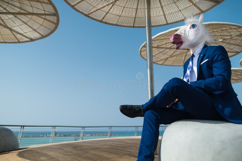 Young man in elegant suit sits under stylish umbrellas on the city waterfront. Unusual businessman in funny mask relaxes on city promenade. Unicorn is enjoying stock images