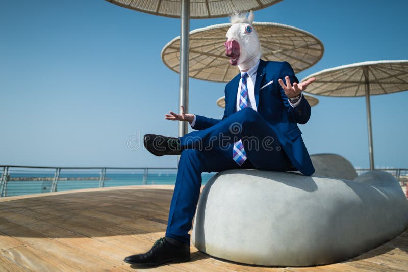 Young man in suit sits under umbrellas on the city waterfront. And active gesticulate. Unusual manager in comical mask spreads his hands on city promenade stock images