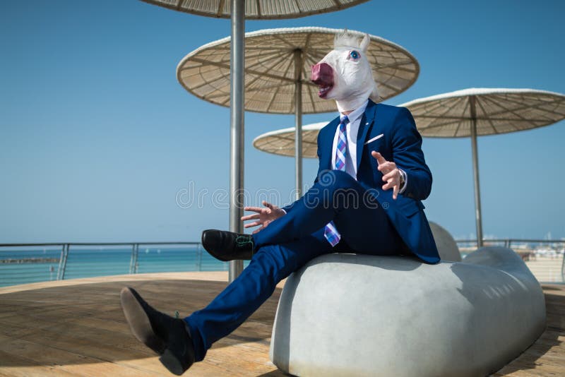 Young manager in suit under umbrellas and spreads his hands. On the city waterfront. Unusual manager in comical mask is surprised. Funny unicorn sits at city royalty free stock photography
